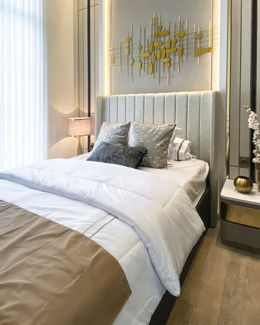 Bedroom with a cream fabric headboard, vertical gold stainless trim on the wall, and a brass line-patterned wall piece