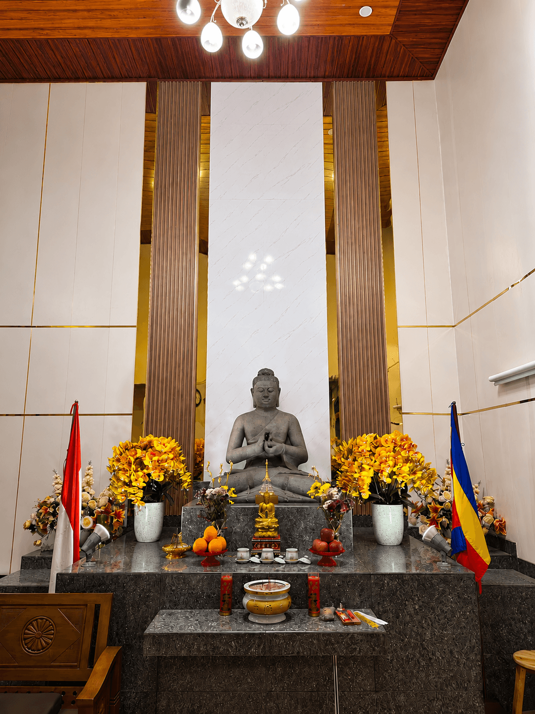 Temple altar under a timber ceiling: white walls lined with gold stainless framing a stone Buddha statue, with yellow flower arrangements on both sides